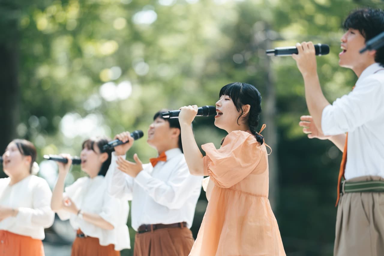 緑の木々を背景に、笑顔で歌うアカペラグループ